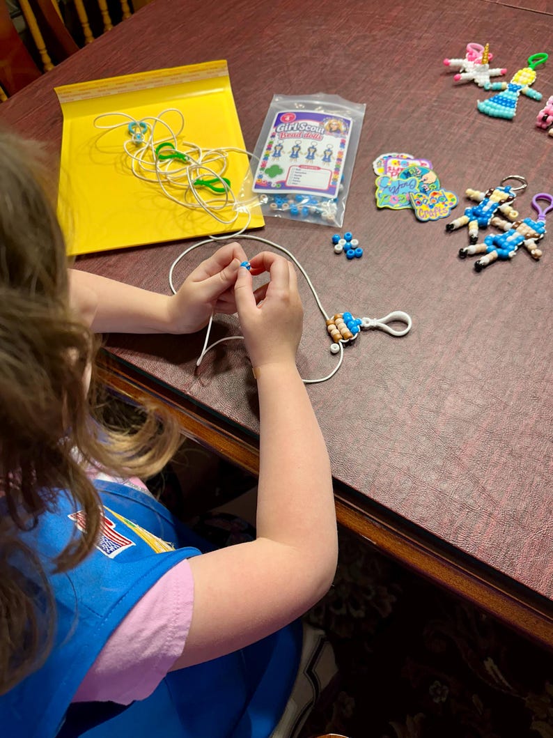 May include: A craft kit for making Girl Scout bead dolls, featuring a clear plastic bag with the text Girl Scout Beadolls, assorted colorful plastic beads, white cord, white lobster clasps, and several finished bead doll keychains.