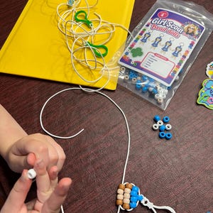 Op de afbeelding: Een Girl Scout Bead Dolls knutselset met een doorzichtige plastic zak met diverse blauwe, witte en beige plastic kralen, wit touw en groene plastic clips, inclusief een instructiekaart met afbeeldingen van de kralenpoppetjes.