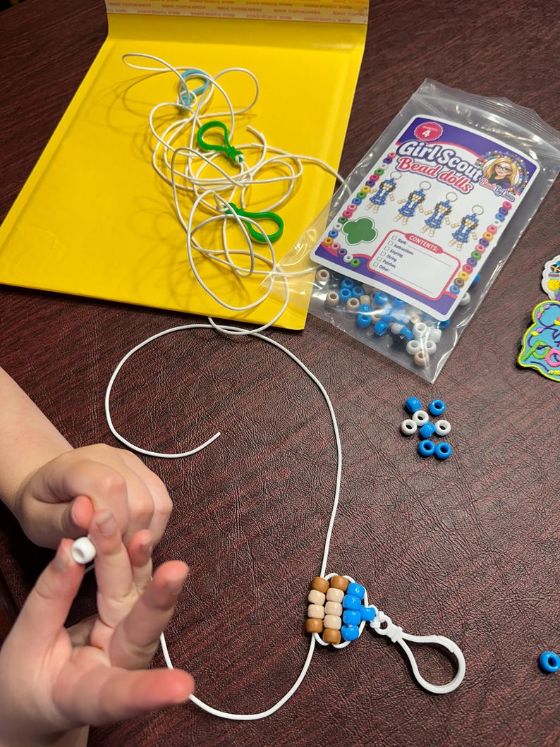 Op de afbeelding: Een Girl Scout Bead Dolls knutselset met een doorzichtige plastic zak met diverse blauwe, witte en beige plastic kralen, wit touw en groene plastic clips, inclusief een instructiekaart met afbeeldingen van de kralenpoppetjes.