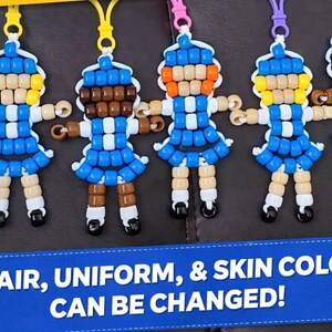 May include: A collection of five small doll-shaped keychains crafted from colorful plastic pony beads, each featuring a blue dress, white accents, and a plastic clip. A blue banner at the bottom displays the text: HAIR, UNIFORM, & SKIN COLOR CAN BE CHANGED!