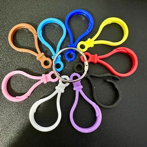 Puede incluir: Una colecci&oacute;n de nueve cierres de mosquet&oacute;n de pl&aacute;stico en varios colores, incluyendo azul, amarillo, rojo, negro, morado, blanco, rosa y marr&oacute;n, todos unidos a una sola anilla de metal plateado dispuesta en un patr&oacute;n circular.