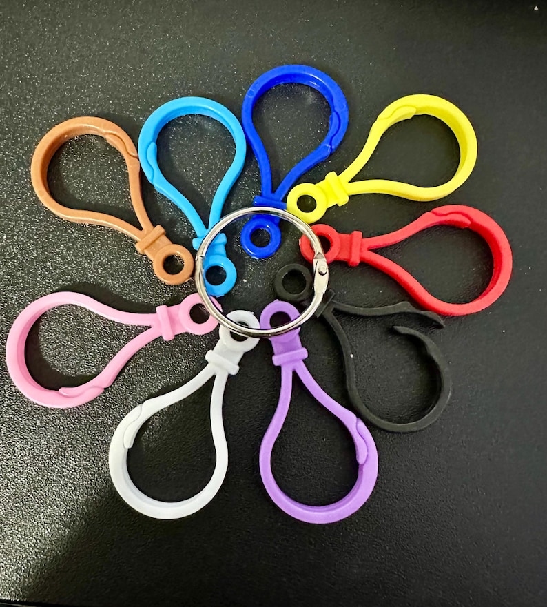 Puede incluir: Una colecci&oacute;n de nueve cierres de mosquet&oacute;n de pl&aacute;stico en varios colores, incluyendo azul, amarillo, rojo, negro, morado, blanco, rosa y marr&oacute;n, todos unidos a una sola anilla de metal plateado dispuesta en un patr&oacute;n circular.