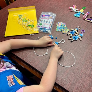 Op de afbeelding: Een knutselset met een doorzichtige plastic zak met het label Girl Scout Bead-dells, met blauwe en witte kralen, wit koord en verschillende afgewerkte kralenpop-sleutelhangers in diverse kleuren, waaronder blauw, roze en wit.