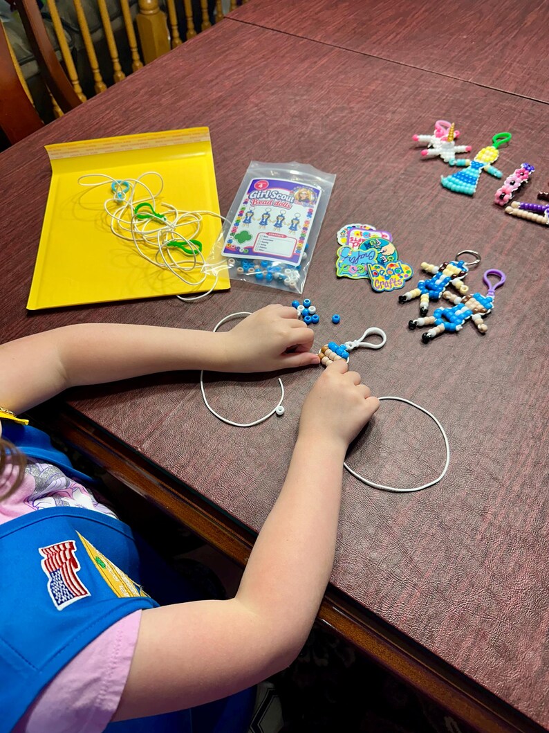 Op de afbeelding: Een knutselset met een doorzichtige plastic zak met het label Girl Scout Bead-dells, met blauwe en witte kralen, wit koord en verschillende afgewerkte kralenpop-sleutelhangers in diverse kleuren, waaronder blauw, roze en wit.