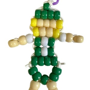 Puede incluir: Un llavero con forma de figura humana construido con cuentas de pl&aacute;stico en colores verde, amarillo, blanco y negro. La figura est&aacute; ensartada en un cord&oacute;n blanco y cuenta con un cierre de mosquet&oacute;n de pl&aacute;stico morado para colgar en bolsos o llaves.