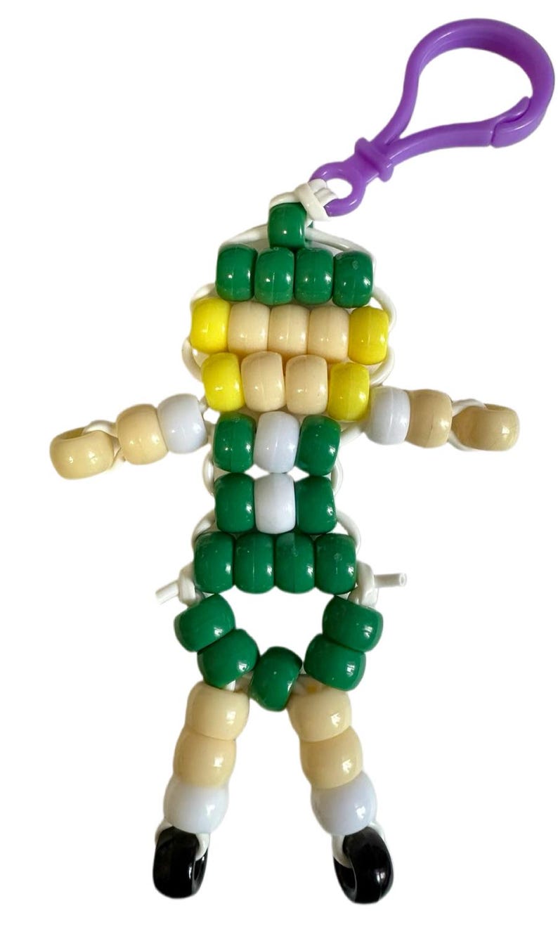 Puede incluir: Un llavero con forma de figura humana construido con cuentas de pl&aacute;stico en colores verde, amarillo, blanco y negro. La figura est&aacute; ensartada en un cord&oacute;n blanco y cuenta con un cierre de mosquet&oacute;n de pl&aacute;stico morado para colgar en bolsos o llaves.
