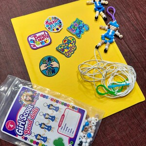 Op de afbeelding: Een knutselset met vier kralenpoppetje-sleutelhangers, vijf geborduurde patches met tekst zoals Bead Fun en Bead Crafts, een rol wit touw met groene clips en een plastic zakje met diverse kralen en een Girl Scout Bead Dolls label.