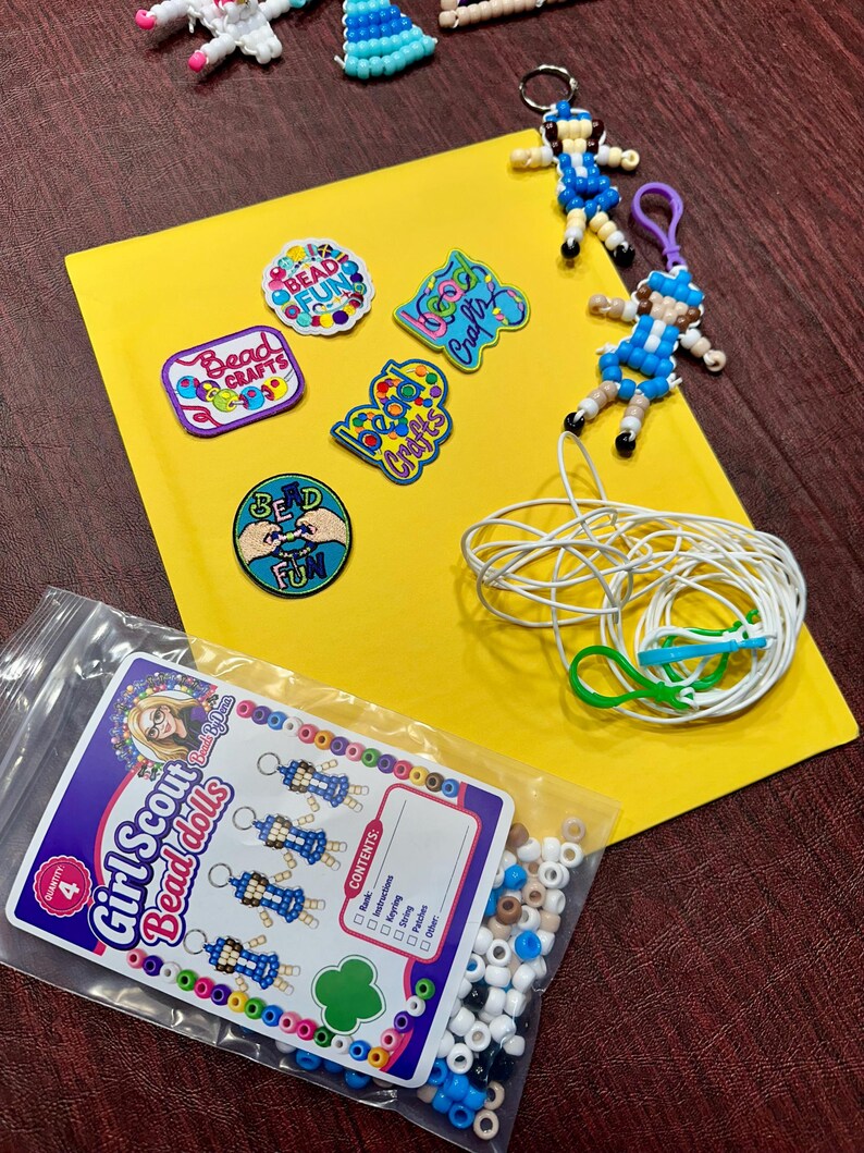 Op de afbeelding: Een knutselset met vier kralenpoppetje-sleutelhangers, vijf geborduurde patches met tekst zoals Bead Fun en Bead Crafts, een rol wit touw met groene clips en een plastic zakje met diverse kralen en een Girl Scout Bead Dolls label.