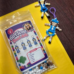 Op de afbeelding: Een doorzichtige plastic zak met een set voor het maken van vier kralenpoppen, gelabeld Girl Scout Bead dolls, met een inhoudslijst inclusief rang, instructies, sleutelhanger, touw en patches. Twee voltooide kralenpoppen-sleutelhangers liggen ernaast.