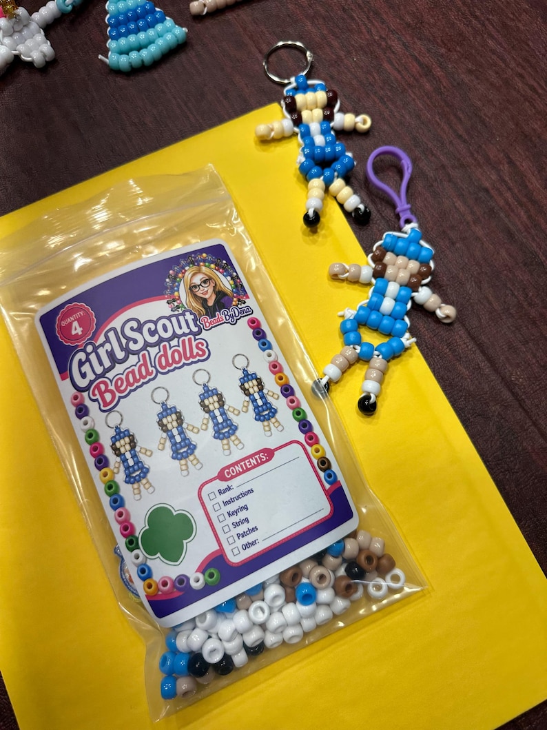 Op de afbeelding: Een doorzichtige plastic zak met een set voor het maken van vier kralenpoppen, gelabeld Girl Scout Bead dolls, met een inhoudslijst inclusief rang, instructies, sleutelhanger, touw en patches. Twee voltooide kralenpoppen-sleutelhangers liggen ernaast.