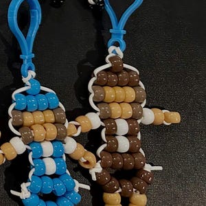 Puede incluir: Dos llaveros hechos a mano con cuentas de pl&aacute;stico en forma de peque&ntilde;as figuras. Una figura presenta cuentas azules, blancas y color canela, mientras que la otra usa cuentas marrones, canela y blancas. Ambos incluyen clips azules de pl&aacute;stico.