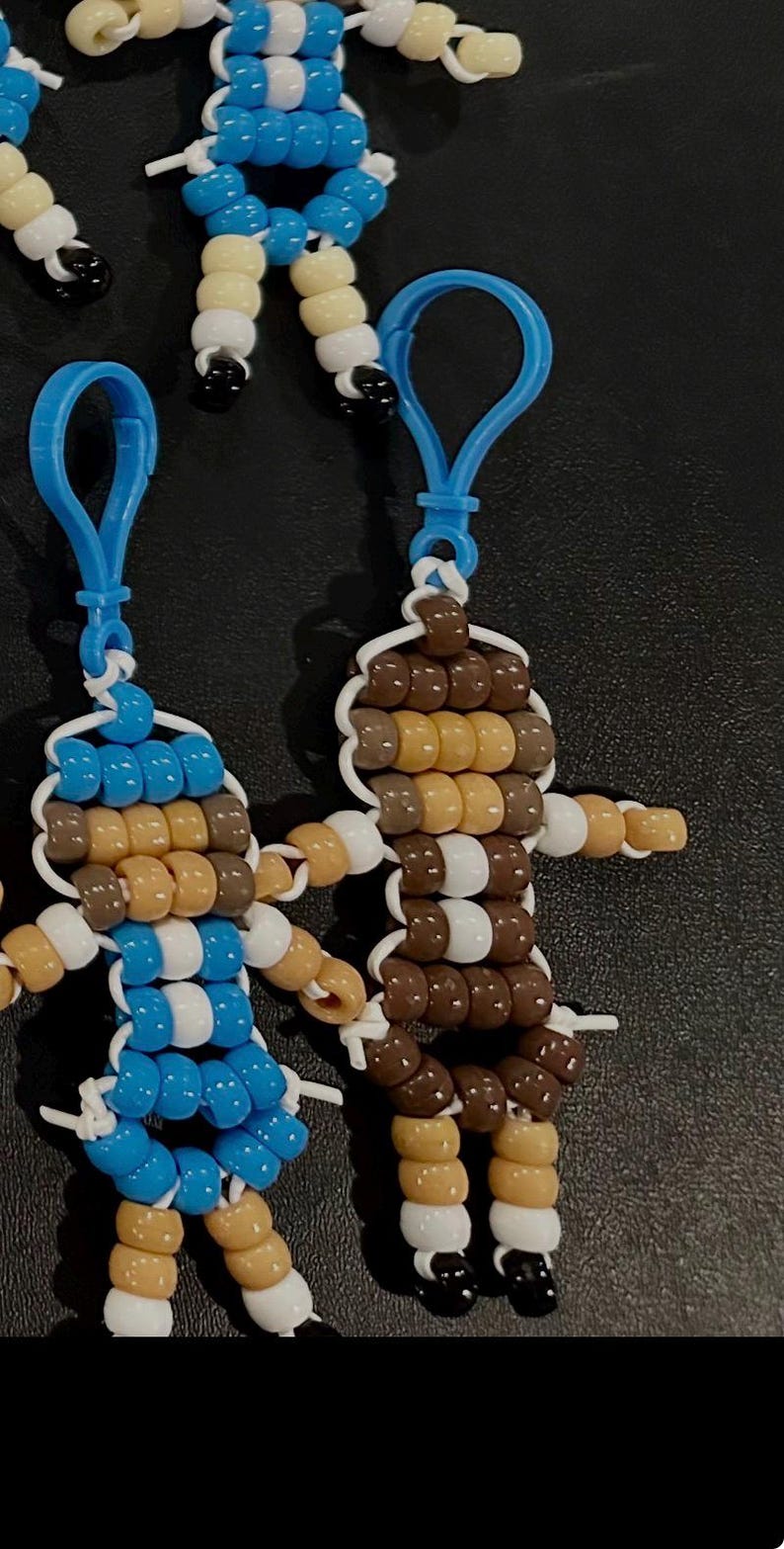 Puede incluir: Dos llaveros hechos a mano con cuentas de pl&aacute;stico en forma de peque&ntilde;as figuras. Una figura presenta cuentas azules, blancas y color canela, mientras que la otra usa cuentas marrones, canela y blancas. Ambos incluyen clips azules de pl&aacute;stico.