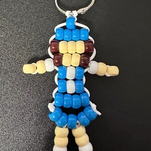 May include: A keychain featuring a small figure constructed from plastic pony beads in blue, white, cream, and dark brown. The figure has a round head, outstretched arms, and two legs, all strung together on a white cord with a silver metal ring.
