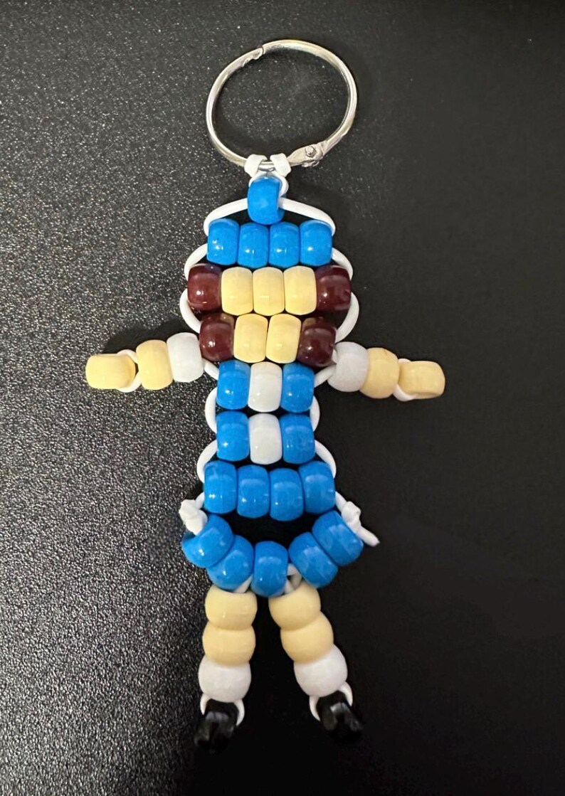 May include: A keychain featuring a small figure constructed from plastic pony beads in blue, white, cream, and dark brown. The figure has a round head, outstretched arms, and two legs, all strung together on a white cord with a silver metal ring.