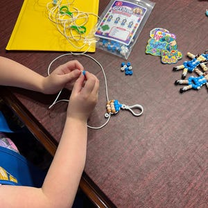 Puede incluir: Un kit de manualidades de mu&ntilde;ecas de cuentas Girl Scout que incluye una bolsa de pl&aacute;stico transparente con cuentas azules, blancas y beige, un cord&oacute;n blanco, un mosquet&oacute;n de pl&aacute;stico blanco, una tarjeta de instrucciones y llaveros terminados.