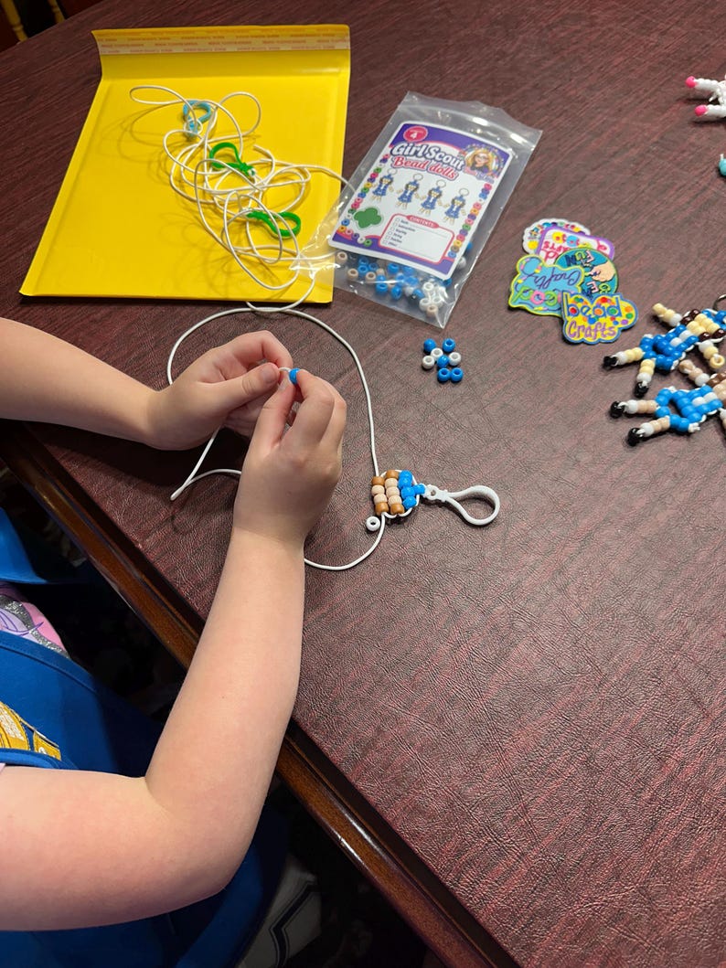 Puede incluir: Un kit de manualidades de mu&ntilde;ecas de cuentas Girl Scout que incluye una bolsa de pl&aacute;stico transparente con cuentas azules, blancas y beige, un cord&oacute;n blanco, un mosquet&oacute;n de pl&aacute;stico blanco, una tarjeta de instrucciones y llaveros terminados.