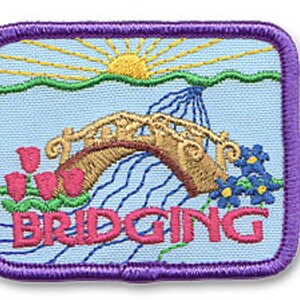 Puede incluir: Un parche bordado rectangular con borde morado sobre fondo azul claro, que muestra un sol amarillo, un puente arqueado color canela sobre agua azul, tulipanes rosas y flores azules. La palabra BRIDGING est&aacute; bordada en letras rosas abajo.