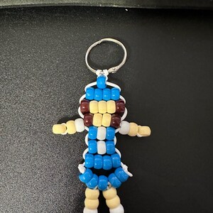 Puede incluir: Un llavero hecho de cuentas de pl&aacute;stico con forma de figura humana. La figura presenta un torso azul y blanco, brazos y piernas de color amarillo claro, una cabeza azul y amarilla con detalles marrones y pies peque&ntilde;os de color negro, unidos por un cord&oacute;n blanco.