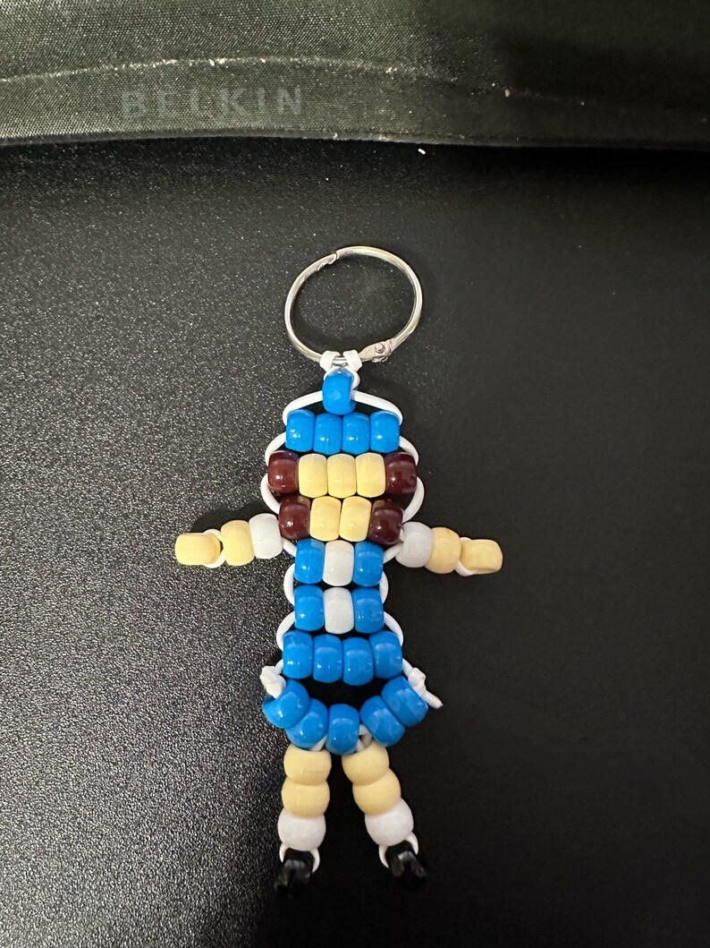 Puede incluir: Un llavero hecho de cuentas de pl&aacute;stico con forma de figura humana. La figura presenta un torso azul y blanco, brazos y piernas de color amarillo claro, una cabeza azul y amarilla con detalles marrones y pies peque&ntilde;os de color negro, unidos por un cord&oacute;n blanco.