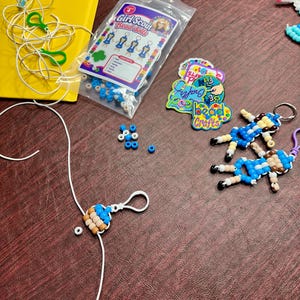 Op de afbeelding: Een knutselset met een doorzichtige plastic zak met het label Girl Scout Bead dolls, met blauwe, witte en beige kralen. Inbegrepen zijn twee afgewerkte kralenpoppetje-sleutelhangers, losse blauwe kralen, wit koord, groene plastic clips en ronde stickers met de tekst bead Crafts.