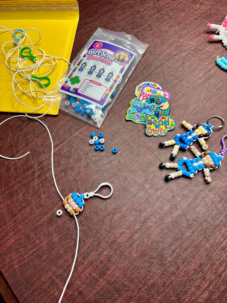 Op de afbeelding: Een knutselset met een doorzichtige plastic zak met het label Girl Scout Bead dolls, met blauwe, witte en beige kralen. Inbegrepen zijn twee afgewerkte kralenpoppetje-sleutelhangers, losse blauwe kralen, wit koord, groene plastic clips en ronde stickers met de tekst bead Crafts.