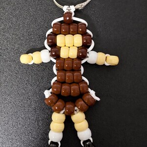 Puede incluir: Un llavero con una figura hecha de cuentas de pl&aacute;stico en color marr&oacute;n, crema, blanco y negro. La figura tiene una cabeza redonda, brazos extendidos y dos piernas, todo unido con un cord&oacute;n blanco y sujeto a una anilla de metal plateado.