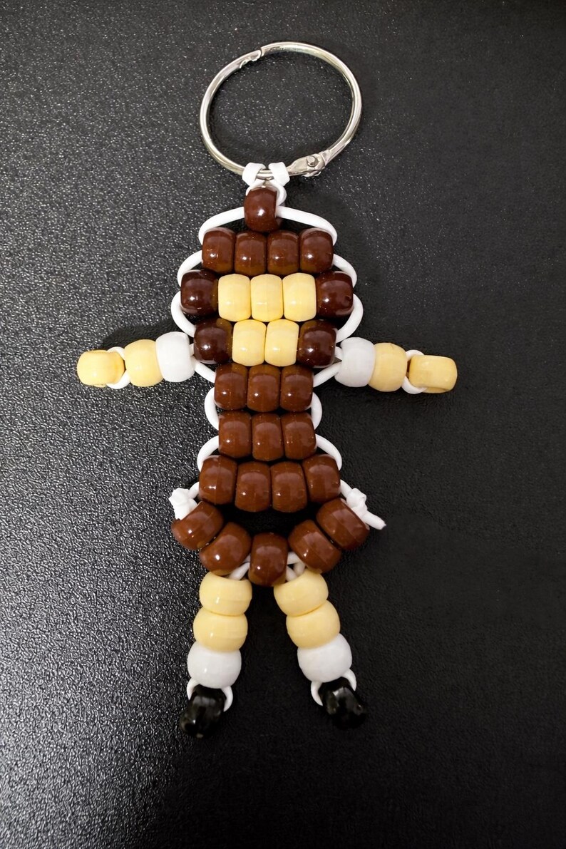 Puede incluir: Un llavero con una figura hecha de cuentas de pl&aacute;stico en color marr&oacute;n, crema, blanco y negro. La figura tiene una cabeza redonda, brazos extendidos y dos piernas, todo unido con un cord&oacute;n blanco y sujeto a una anilla de metal plateado.