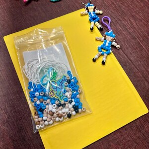 May include: A clear plastic bag containing white, blue, and tan pony beads with white elastic cords and a small green charm. Two finished bead keychains shaped like figures in blue and tan outfits are placed next to the bag on a yellow surface.