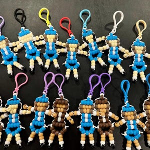 Puede incluir: Una colecci&oacute;n de quince llaveros hechos a mano con cuentas de pl&aacute;stico, cada uno con forma de peque&ntilde;a figura humana. Las figuras presentan varias combinaciones de colores en azul, blanco, marr&oacute;n y beige, con bucles de pl&aacute;stico para su sujeci&oacute;n.
