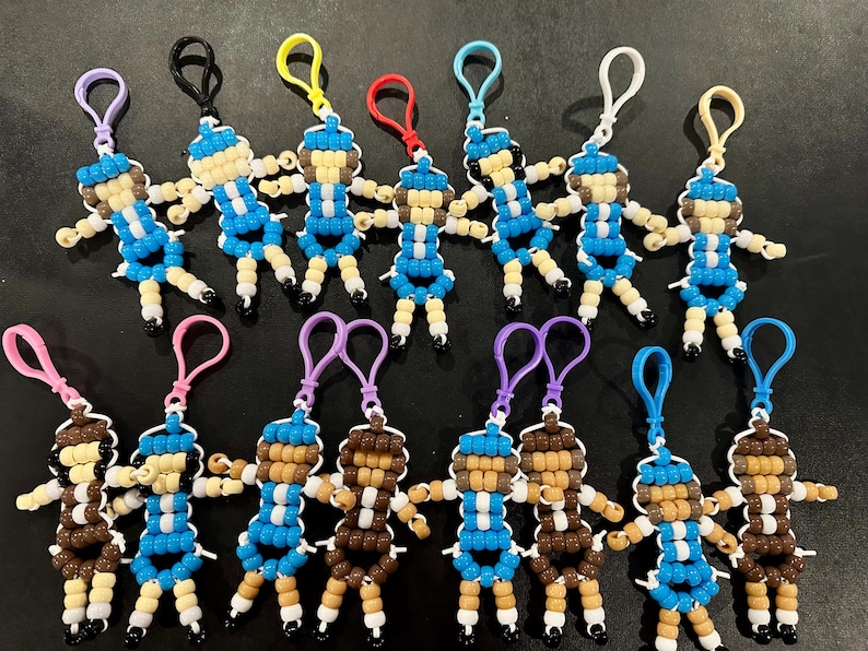 Puede incluir: Una colecci&oacute;n de quince llaveros hechos a mano con cuentas de pl&aacute;stico, cada uno con forma de peque&ntilde;a figura humana. Las figuras presentan varias combinaciones de colores en azul, blanco, marr&oacute;n y beige, con bucles de pl&aacute;stico para su sujeci&oacute;n.
