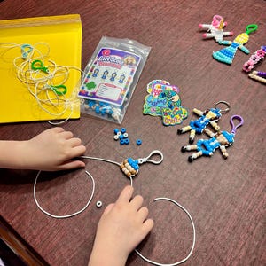 May include: A Girl Scout Bead Dolls craft kit featuring a plastic bag of assorted beads, a printed instruction card, several finished doll keychains in various colors, loose white cords, and colorful stickers on a dark wooden surface.
