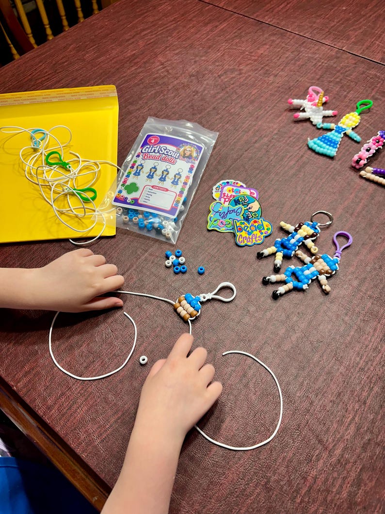 May include: A Girl Scout Bead Dolls craft kit featuring a plastic bag of assorted beads, a printed instruction card, several finished doll keychains in various colors, loose white cords, and colorful stickers on a dark wooden surface.