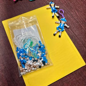Op de afbeelding: Een doorzichtig plastic zakje met witte, blauwe en beige pony-kralen, witte elastische koorden en een kleine groene bedel. Twee afgewerkte kralen-sleutelhangers in de vorm van figuren met blauwe en beige outfits liggen naast het zakje.