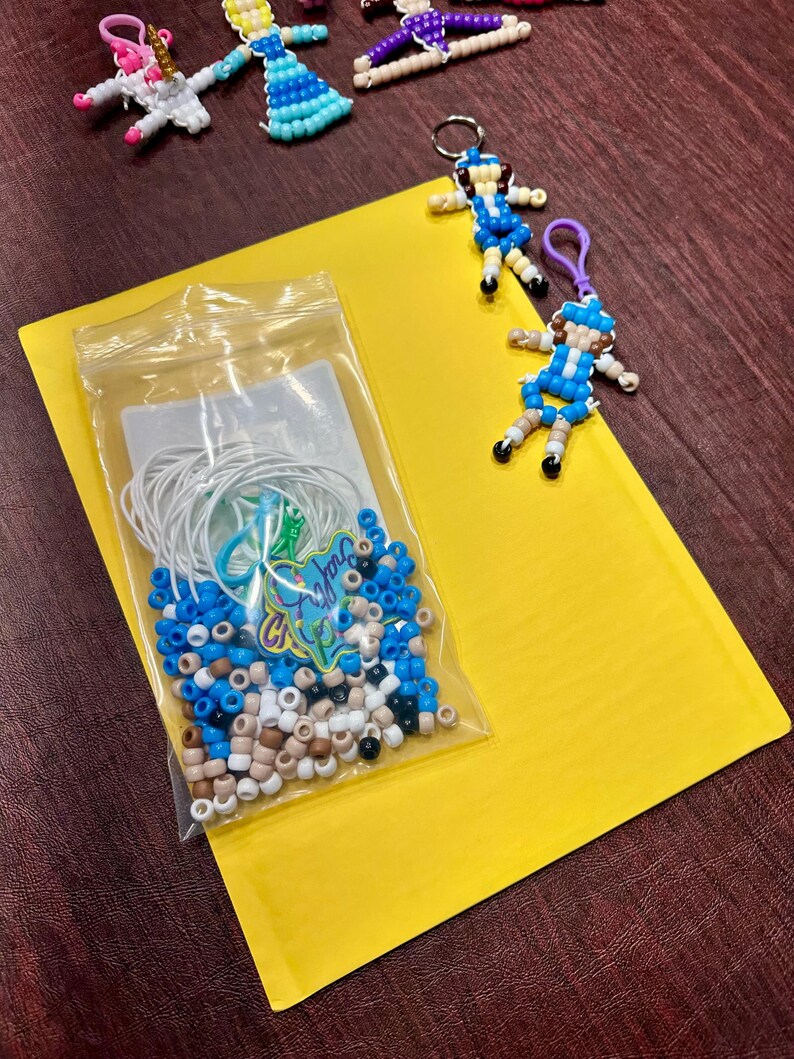 Op de afbeelding: Een doorzichtig plastic zakje met witte, blauwe en beige pony-kralen, witte elastische koorden en een kleine groene bedel. Twee afgewerkte kralen-sleutelhangers in de vorm van figuren met blauwe en beige outfits liggen naast het zakje.