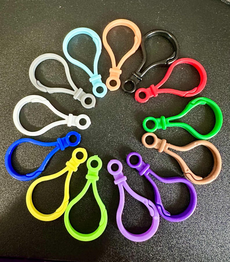 May include: A collection of twelve plastic lobster claw clasps arranged in a circular pattern. The clasps are available in various solid colors including black, white, gray, red, orange, yellow, light blue, green, lime green, purple, blue, and tan.