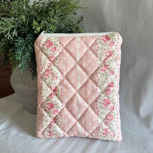 May include: A quilted pouch with a white zipper, featuring a pink diamond pattern and floral accents. The pouch has a soft, padded appearance, with a decorative floral border. The overall design is a mix of pink, white, and floral patterns.