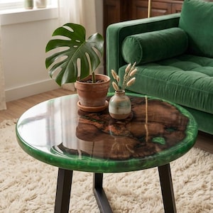 Puede incluir: Una mesa de centro redonda con un centro de losa de madera natural rodeado por un borde de resina verde translúcida y brillante. La mesa se apoya sobre una base de metal negro de tres patas. Una maceta de terracota con una planta monstera y un pequeño jarrón descansan encima.