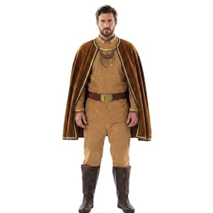 May include: A tan long-sleeved tunic and matching trousers paired with a dark brown velvet cape featuring gold trim. The outfit includes a brown belt with a gold buckle, a gold-toned necklace with dangling pendants, and dark brown leather boots.