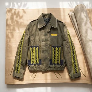 Puede incluir: Chaqueta verde oliva con cuello, cierre de botones y múltiples bolsillos. La chaqueta presenta detalles decorativos amarillos y negros en las mangas y el frente, con un pequeño parche amarillo en el pecho. La chaqueta se exhibe sobre una superficie de madera.