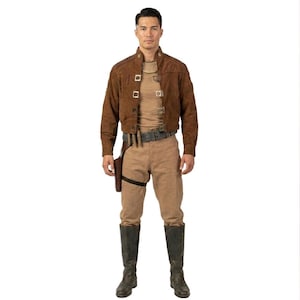Può includere: Una giacca color marrone effetto camoscio con colletto alto e chiusure metalliche quadrate, indossata sopra una maglia beige e pantaloni marrone chiaro. L'abbigliamento include una cintura scura con fondina e stivali alti grigio scuro al polpaccio.