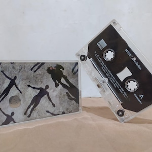 Puede incluir: Dos cintas de cassette transparentes. Una tiene una etiqueta negra con texto blanco que dice "MUSE / Absolution". La otra tiene una portada con fondo gris y siluetas negras. Las carcasas de los casetes están sobre una superficie beige.