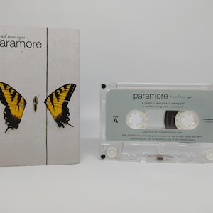 May include: A cassette tape and album cover for the band Paramore's album "Brand New Eyes." The album cover features a butterfly design with yellow and black wings. The cassette tape is clear with a gray label.