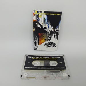 May include: A cassette tape and its cover for the film "The Fast and the Furious: Tokyo Drift." The cover shows a black car with orange flames and the film title. The cassette tape is transparent with black text.