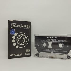 May include: A black cassette tape case and cassette tape for the band Blink-182. The case has the band's logo and the words "Greatest Hits". The cassette tape is clear with black text.