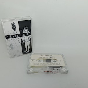 May include: A clear plastic cassette case featuring the album cover for Death Grips The Money Store, displaying a black and white illustration of two figures. Beside it lies a clear cassette tape with a white label printed with the text Epic.