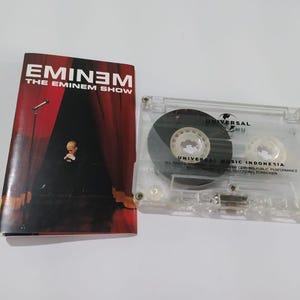 Puede incluir: Una cinta de cassette y la portada del álbum "The Eminem Show" de Eminem. La portada del álbum presenta el título en texto blanco y rojo, con una escena de escenario y una figura en el escenario. La cinta de cassette es transparente.