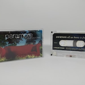 May include: A clear cassette tape case and cassette featuring the band Paramore's album "all we know is falling." The album art shows a red couch in a grassy area. The cassette tape has the album title and band name printed on it.