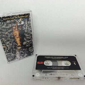 Könnte beinhalten: Eine transparente Kunststoff-Kassettenhülle mit dem Albumcover von Siouxsie and the Banshees Juju mit einer dunklen, abstrakten Grafik, neben einer passenden Kassette mit schwarzem Etikett, weißem Text und der aufgedruckten Nummer 1.