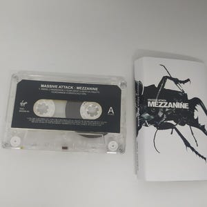 May include: A clear cassette tape and album cover for Massive Attack's "Mezzanine." The cassette tape has a black label with white text. The album cover is white with a black insect graphic and the album title.