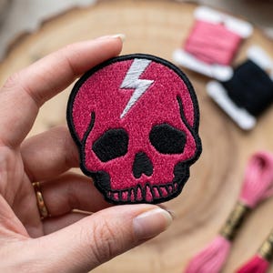 Könnte beinhalten: Ein pinkfarbener Totenkopf-Aufnäher mit einem weißen Blitzdesign. Der Schädel hat schwarze Augenhöhlen und eine schwarze Umrandung. Der Aufnäher wird in einer Hand gehalten, mit Garnrollen im Hintergrund.
